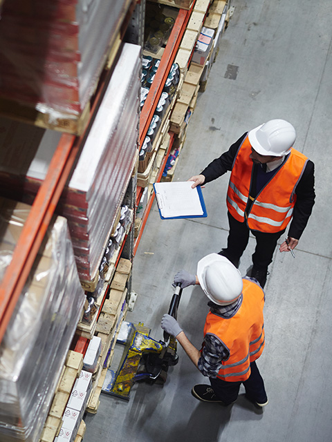 two warehouse workers inspecting goods in a storage facility wearing safety vests and helmets while checking an inventory list for twelve specific items