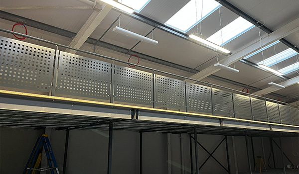 A view of an industrial space featuring a metal railing with perforations on an upper level, supported by a steel structure, under bright ceiling lights and skylights, showcasing four distinct sections of the railing.